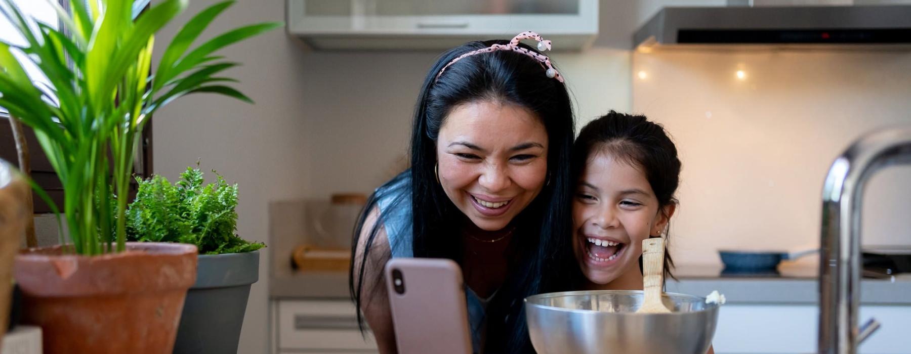 mother and young daughter smile at their phone as they cook in the kitchen